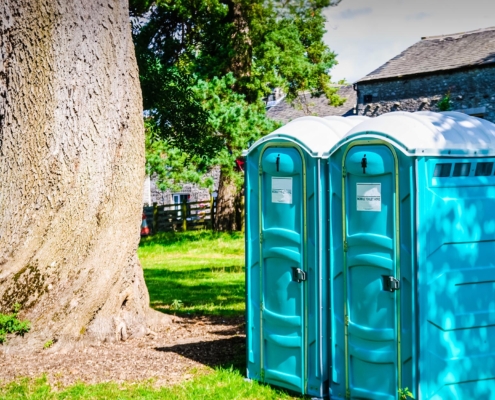 Side view of two porta potties sitting next to a large tree